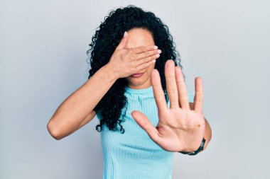 Young hispanic woman with curly hair wearing casual blue t shirt covering eyes with hands and doing stop gesture with sad and fear expression. embarrassed and negative concept. 
