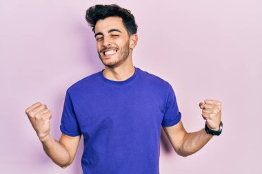 Gündelik tişört giyen genç İspanyol adam çok mutlu ve heyecanlı. Kolları havada, gülümseyen ve başarı için çığlık atan kazanan bir jest yapıyor. kutlama konsepti. 