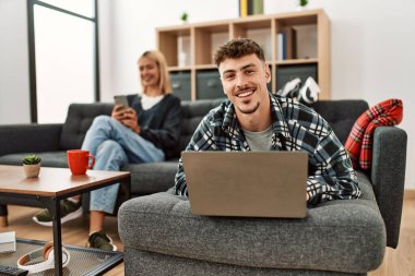 Evdeki kanepede oturan genç beyaz çift dizüstü bilgisayar ve akıllı telefon kullanarak mutlu bir şekilde gülümsüyor..