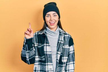 Beautiful woman with blue eyes wearing wool cap smiling with an idea or question pointing finger up with happy face, number one 