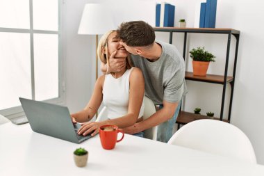 Genç, beyaz bir çift evdeki masada dizüstü bilgisayarla sarılıp öpüşüyor..