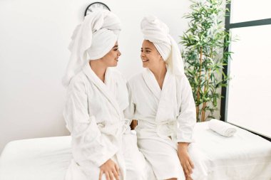 Bornoz giymiş bir çift birbirlerine sarılıyorlar güzellik merkezinde masaj masasında oturuyorlar.