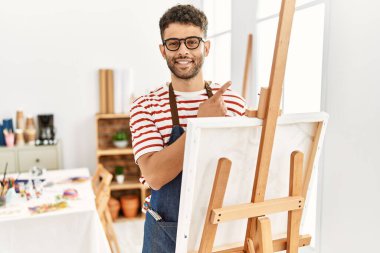 Sanat stüdyosundaki Arap genç adam, elini kenarı işaret ederek reklamını yapıyor, ciddi ve sakin bir yüz ifadesi. 