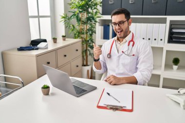 Sakallı genç İspanyol doktor klinikte online randevu veriyor. Başparmağıyla yan tarafa işaret ediyor. Ağzı açık gülümsüyor. 