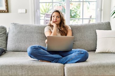 Güzel, genç, esmer bir kadın evdeki bilgisayarı kullanarak kanepede oturuyor. Elleri çenesinde soru sorarken, düşünceli bir ifade takınıyor. Düşünceli bir yüzle gülümsüyor. şüphe kavramı. 