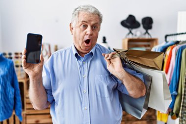 Gri saçlı, alışveriş torbası tutan ve şok olmuş suratıyla akıllı telefon ekranı gösteren son sınıf öğrencisi şüpheci ve alaycı görünüyor, ağzı açık bir şekilde şaşırmış. 