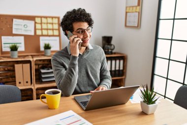 Genç İspanyol iş adamı akıllı telefonla konuşuyor ve ofiste dizüstü bilgisayar kullanıyor..