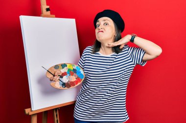 Middle age hispanic woman standing drawing with palette by painter easel stand cutting throat with hand as knife, threaten aggression with furious violence 