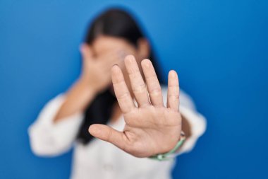 Mavi arka planda duran genç İspanyol kadın elleriyle gözlerini kapatıyor ve üzgün ve korku ifadesiyle dur hareketi yapıyor. utangaç ve olumsuz kavram. 