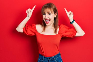 Kırmızı tişörtlü kızıl saçlı genç kadın şaşkın şaşkın ve şaşırmış bir şekilde parmaklarını kaldırıp kollarını kaldırdı.. 