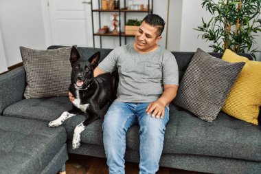 Genç İspanyol adam gülümsüyor. Kanepede köpeğiyle oturuyor..