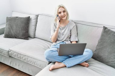 Beyaz bir kadın akıllı telefondan evde dizüstü bilgisayar kullanarak konuşuyor.