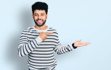 Sakallı, sıradan çizgili süveter giyen genç Arap adam kameraya hayret ediyor ve gülümsüyor.. 