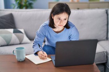 Down sendromlu genç bir kadın kanepede oturuyor ve not defterine evde ders çalışıyor.