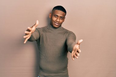 Balıkçı yaka kazak giyen genç siyahi bir adam kameraya sarılmak için gülümseyerek bakıyor. Mutluluğu kucaklayan neşeli bir ifade.. 