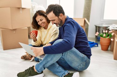 Orta yaşlı İspanyol çift dizüstü bilgisayar kullanıyor ve kahve içiyor. Yeni evinde köpekle yerde oturuyor..