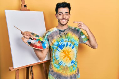 İspanyol asıllı genç adam paletle resim çiziyor, resim sehpası ayakta gülümsüyor ve parmakları, dişleri ve ağzıyla işaret ediyor. diş sağlığı kavramı. 