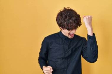 Sarı arka planda gözlük takan genç adam çok mutlu ve heyecanlı. Kolları havada, gülümseyen ve başarı için çığlık atan kazanan bir jest yapıyor. kutlama konsepti. 