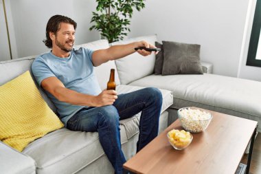 Orta yaşlı beyaz bir adam evde televizyon izlerken kendinden emin gülümsüyor.