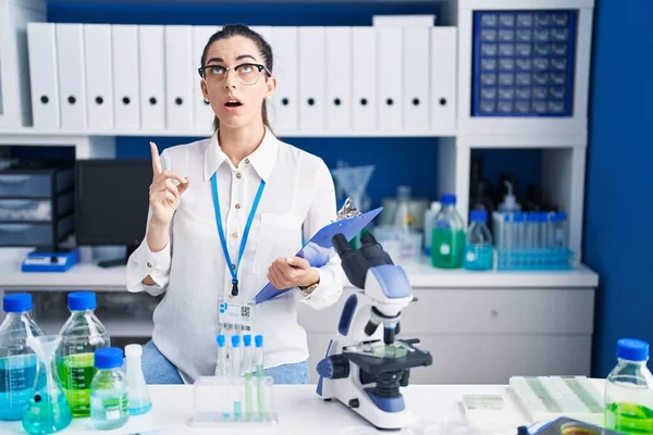 Young brunette woman working at scientist laboratory amazed and surprised looking up and pointing with fingers and raised arms.