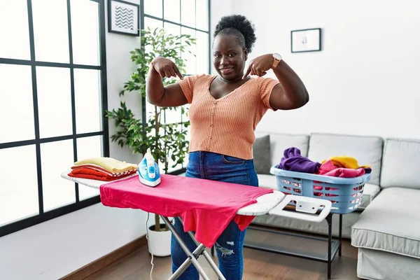 Young african woman ironing clothes at home looking confident with smile on face, pointing oneself with fingers proud and happy.