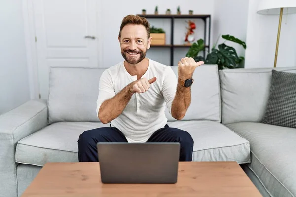 Middle age man using laptop at home pointing to the back behind with hand and thumbs up, smiling confident