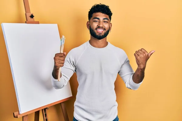 Arab man with beard standing by painter easel stand holding brushes pointing thumb up to the side smiling happy with open mouth