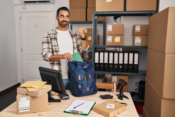 Young hispanic man with beard working at small business ecommerce pointing thumb up to the side smiling happy with open mouth