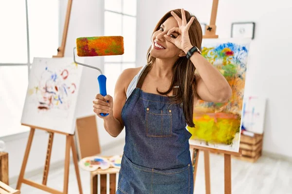 年轻的拉丁艺术家女人拿着粉刷滚筒站在艺术工作室 高兴地微笑着 手拿着手拿着手签 透过手指看 — 图库照片