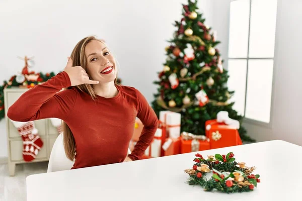 Caucasian young blonde woman sitting on the table by christmas tree smiling doing phone gesture with hand and fingers like talking on the telephone. communicating concepts.