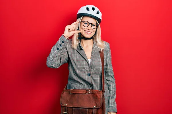 Beautiful caucasian blonde business woman wearing bike helmet smiling and confident gesturing with hand doing small size sign with fingers looking and the camera. measure concept.
