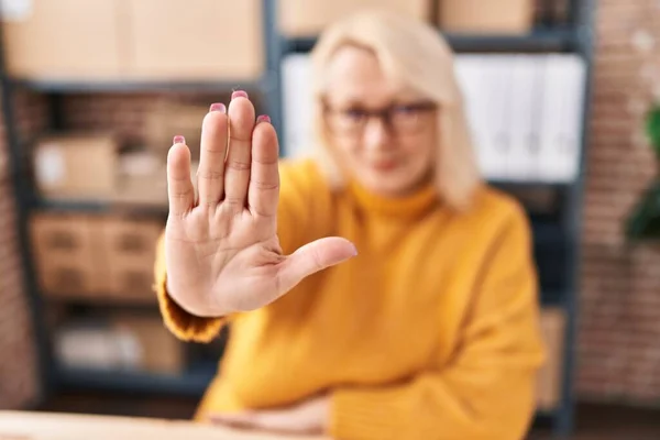 Middle age blonde woman ecommerce business worker doing stop gesture with hands at office