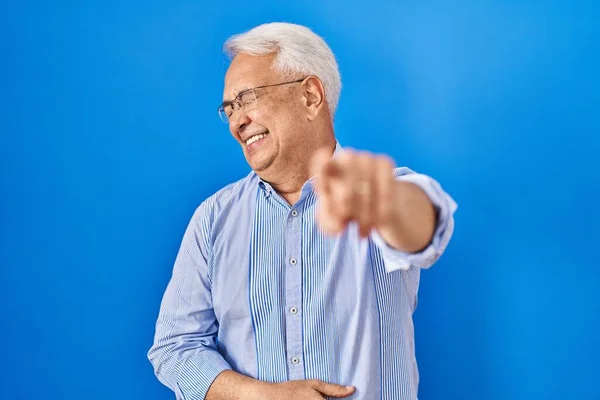 Hispanic senior man wearing glasses laughing at you, pointing finger to the camera with hand over body, shame expression