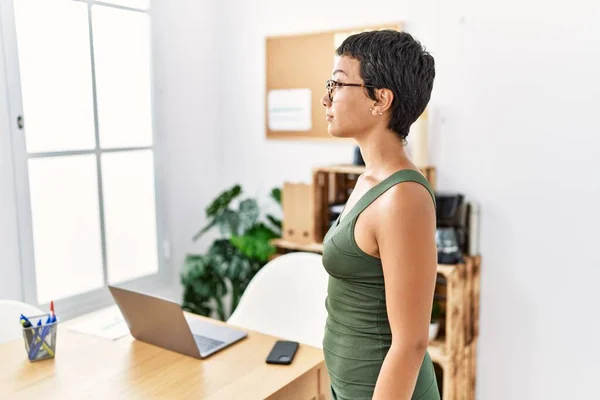 Young hispanic woman with short hair working at the office looking to side, relax profile pose with natural face with confident smile.