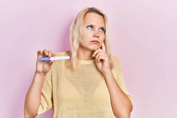 Beautiful caucasian blonde woman holding pregnancy test result serious face thinking about question with hand on chin, thoughtful about confusing idea