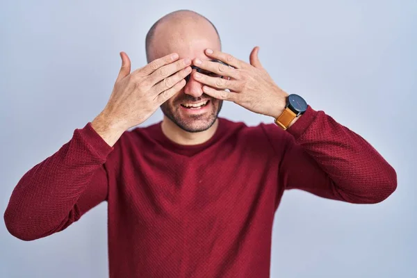 Young bald man with beard standing over white background wearing glasses covering eyes with hands smiling cheerful and funny. blind concept.