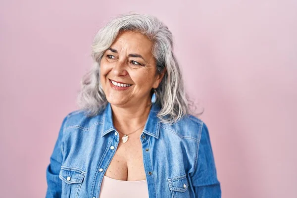 Middle age woman with grey hair standing over pink background looking away to side with smile on face, natural expression. laughing confident.