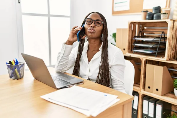 Black woman with braids working at the office speaking on the phone looking at the camera blowing a kiss on air being lovely and sexy. love expression.