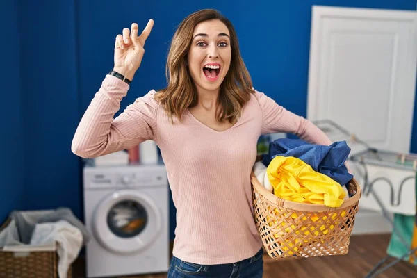Young woman holding laundry basket smiling amazed and surprised and pointing up with fingers and raised arms.