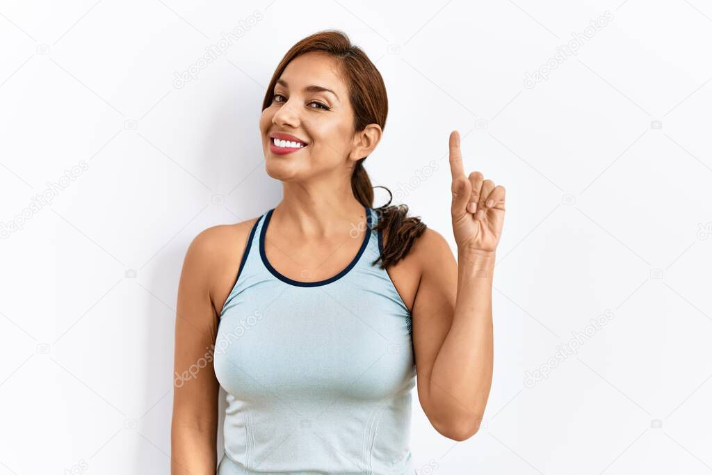 Young latin woman wearing sporty clothes over isolated background showing and pointing up with finger number one while smiling confident and happy. 