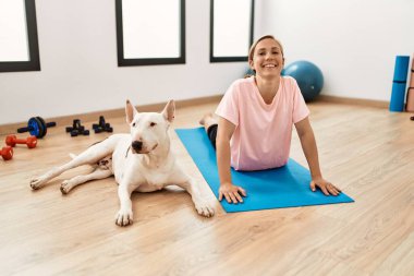 Genç beyaz kadın gülümsüyor spor merkezinde köpekle esniyor.