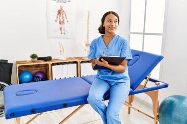 Fizyoterapist üniforması giyen genç Latin bir kadın klinikteki panoya yazı yazıyor.