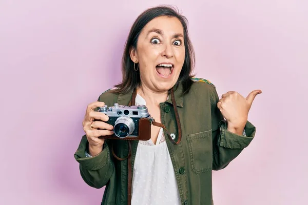 Middle age hispanic woman holding vintage camera pointing thumb up to the side smiling happy with open mouth