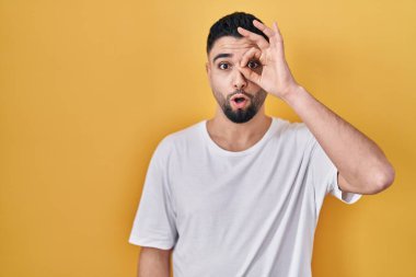 Sarı arka plan üzerine gündelik tişört giyen yakışıklı genç adam şaşırmış yüz ifadesiyle şaşırmış, parmaklarının arasından bakan bir jest yapıyor. İnançsız ifade. 