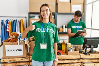 Young blonde girl wearing volunteer t shirt at donation stand doing happy thumbs up gesture with hand. approving expression looking at the camera showing success. 