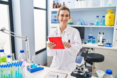 Laboratuvarda dokunmatik ped kullanan bilim adamı üniforması giyen genç bir kadın.