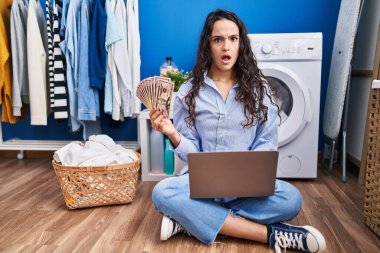 Genç esmer kadın çamaşırlarını yıkıyor. Dizüstü bilgisayarını kullanıyor. Şok olmuş yüzüne para tutuyor. 