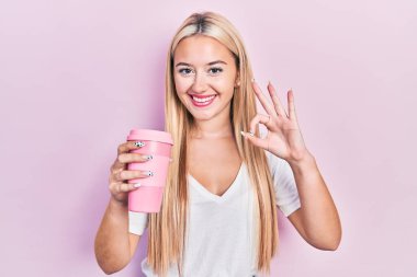 Elinde kahve tutan genç sarışın kız parmaklarıyla imza atıyor, gülümseyerek mükemmel bir sembol oluşturuyor. 