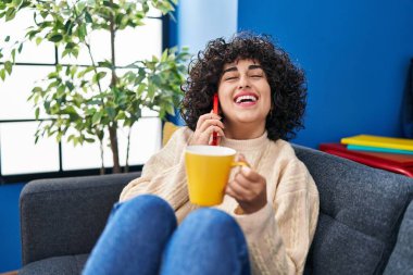 Orta Doğulu genç bir kadın kahve içiyor ve evde akıllı telefondan konuşuyor.