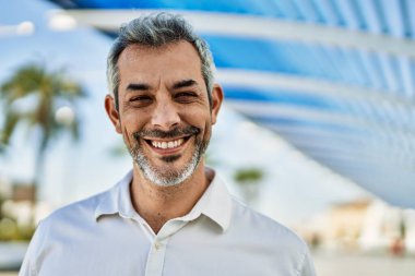 Orta yaşlı, kır saçlı, gülümseyen bir adam şehirde mutlu bir şekilde duruyor..